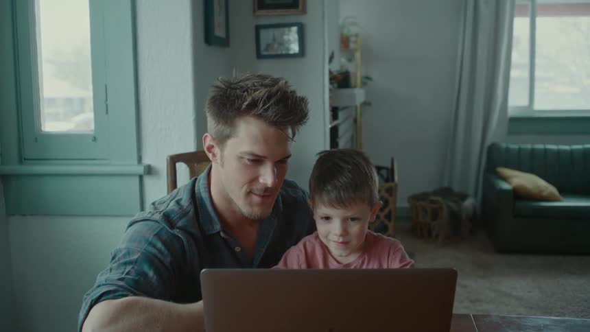 A father sits at his laptop while holding his son on his lap