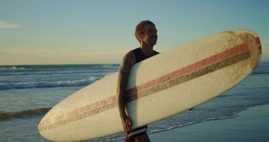 Young tattooed man walks with a longboard on the beach