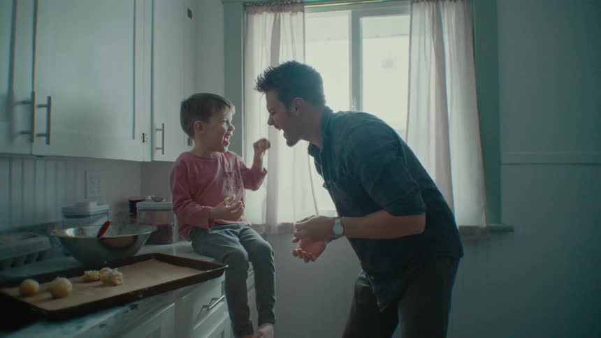 A father smiles while his son sits on the kitchen counter while they make food