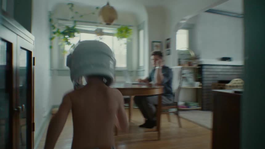 Two boys chase each other inside a house one in an astronauts helmet the other in an animal suit until they pass their father who is on the phone and working on a laptop