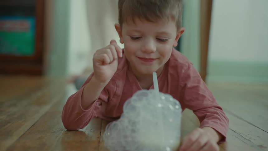 Portrait of a little boy lying on the floor blowing into a cup with a straw to create foam and bubbles