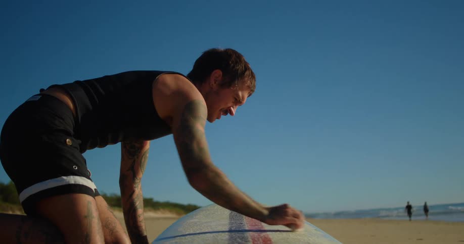 Man sitting on the beach sand applying paraffin on his surfboard