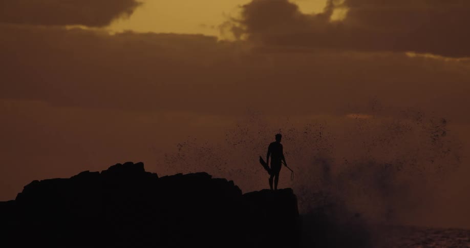 Silhouette of a man on a cliff by the seaside with water splashing upward on a day with a reddish sky