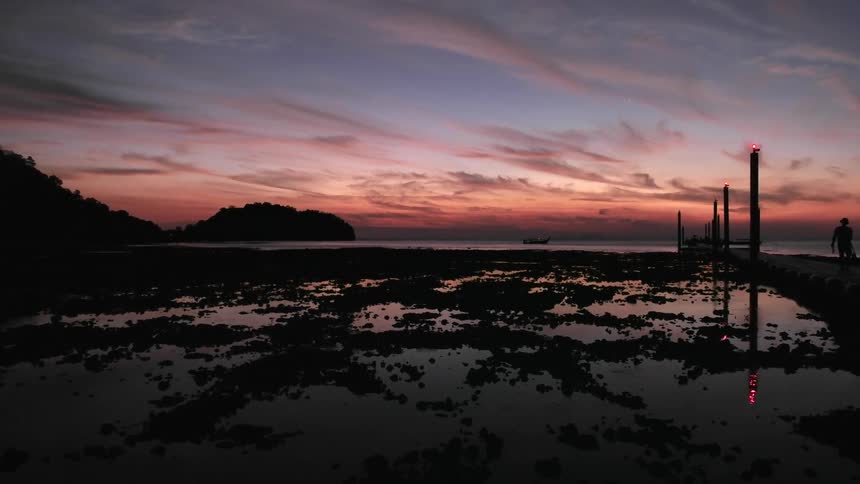 A man walks along a pier beneath a pink and blue cloudy sky