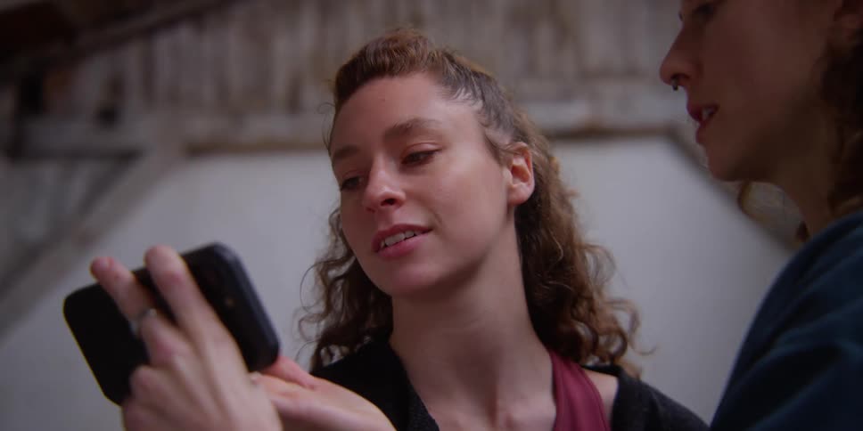Two women talking with one of them showing the other dance moves on a smart phone which the first women tries to repeat