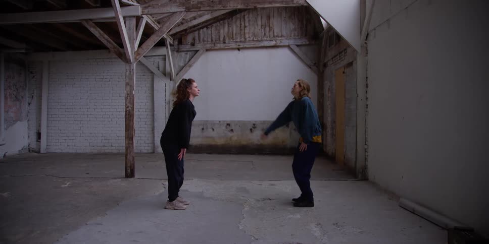 Two standing females stretches their arms and bodies while facing each other under a roof