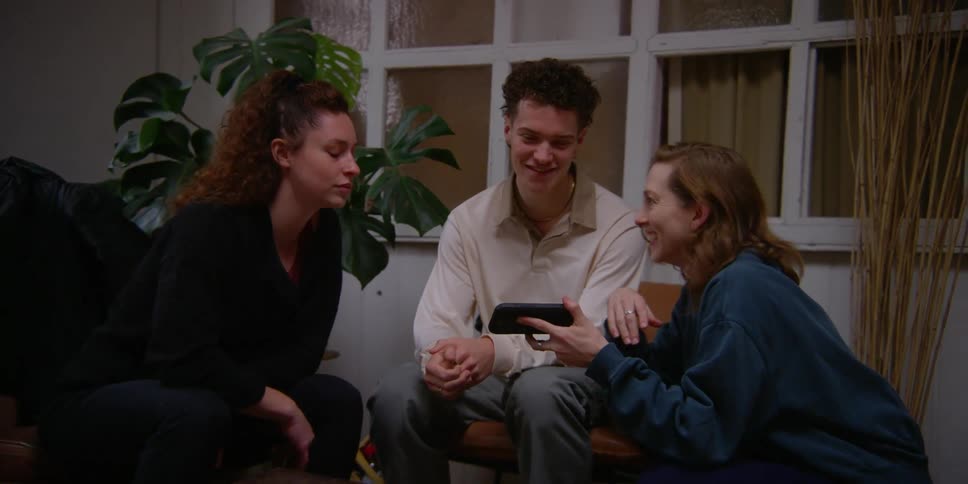 Three people sitting and talking while one of them holds a smart phone at which they point and laugh