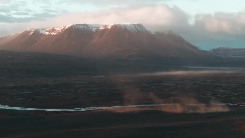 A peaceful landscape with a river on the plains next to a snowy mountain under a cloudy sky