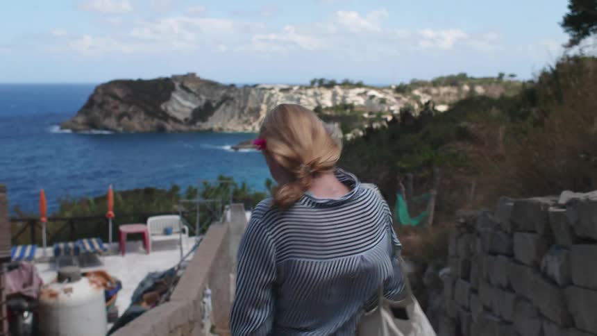 A woman walks downhill near a residence overlooking a coastal cove
