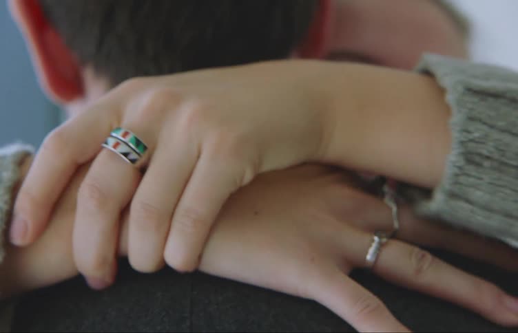 A person hugging a person who has their back turned and their hand has some rings on it