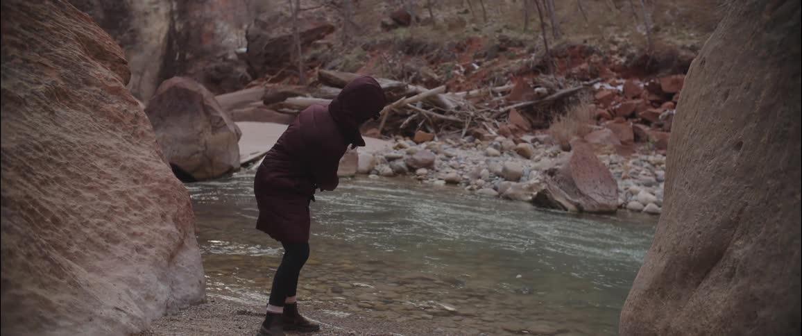 A person in a jacket is throwing a stone into a small stream