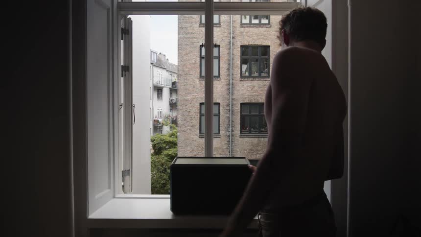 Man without a shirt going towards the window of a building and looking at a black box