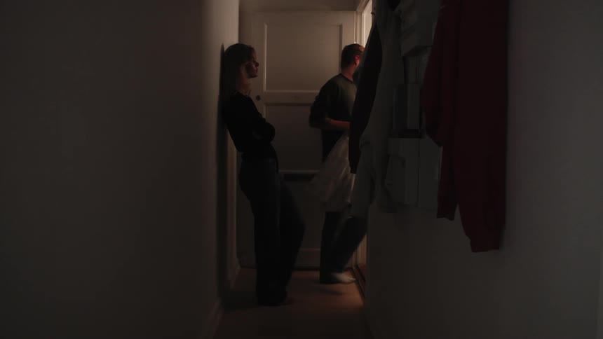 A woman acts sad as a man takes a coat from a hallway and leaves