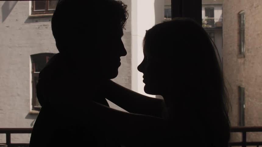 The silhouette of a young man and woman show them embracing in front of a window