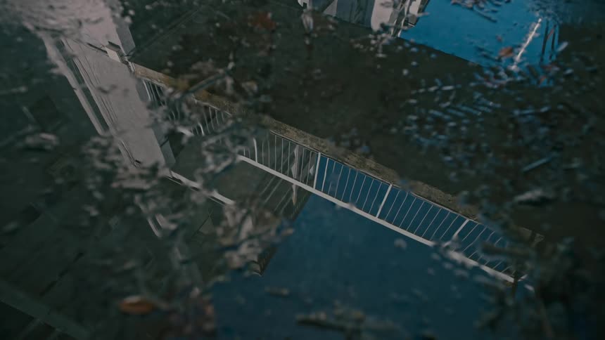 A puddle on the ground reflects a bridge connecting buildings above it