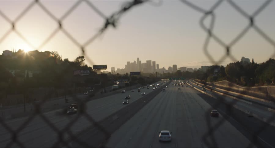 A scenic shot of downtown Los Angeles shot through a hole in a chain linked fence with the freeway running underneath