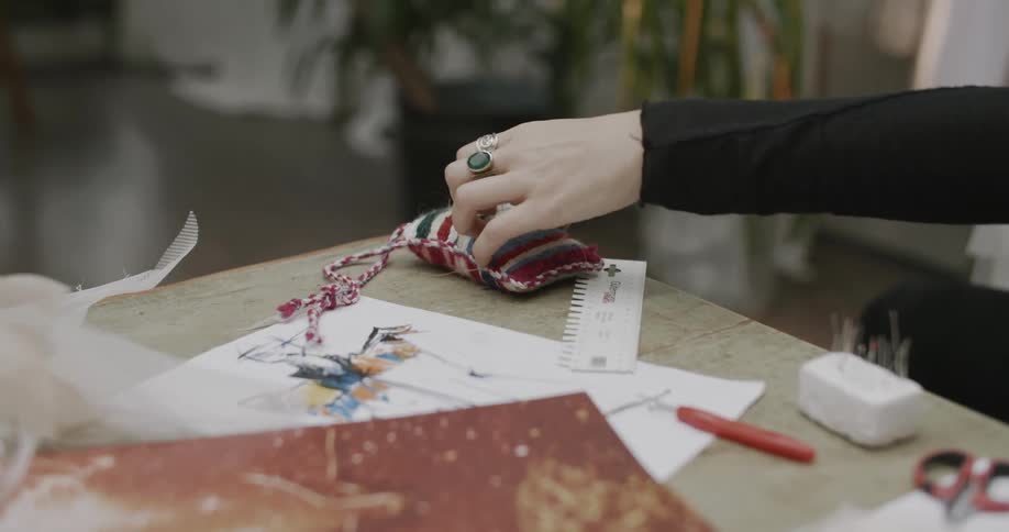 A womans hand takes a pin from a pin cushion on a table next to a sketch on paper