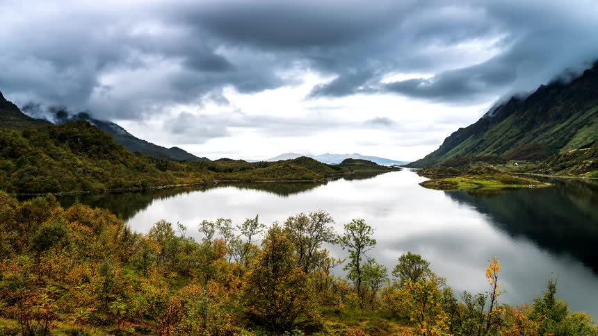 Natural landscape of a reflective lake with mountain ranges around it and heavy clouds passing by
