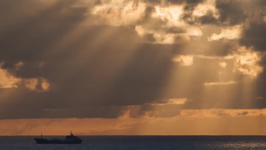 A boat passing at sea in a region with many clouds in the sky that is orange
