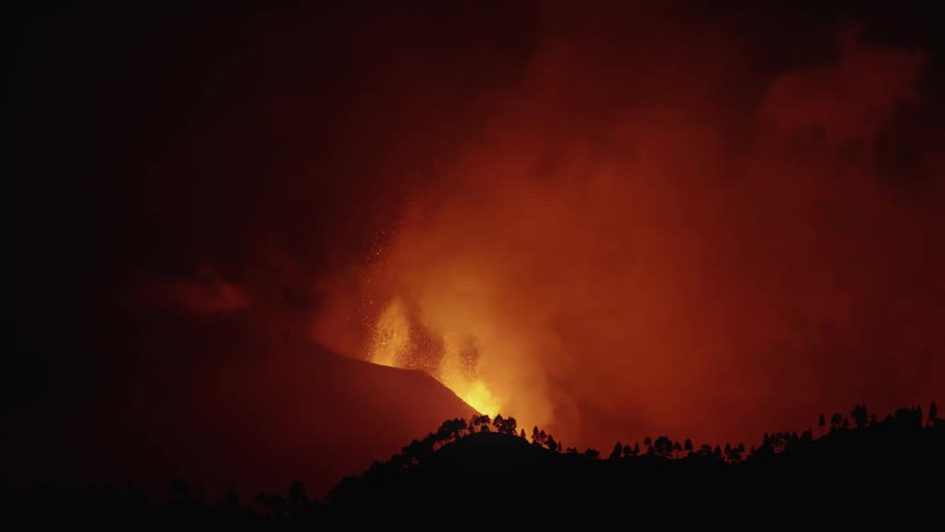 An erupting volcano releasing a lot of lava and smoke upwards and in front of a silhouette of a forest