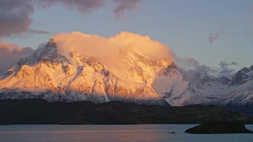 Timelapse of sunrise turning into day with clouds over a snow covered mountain beyond a lakeshore
