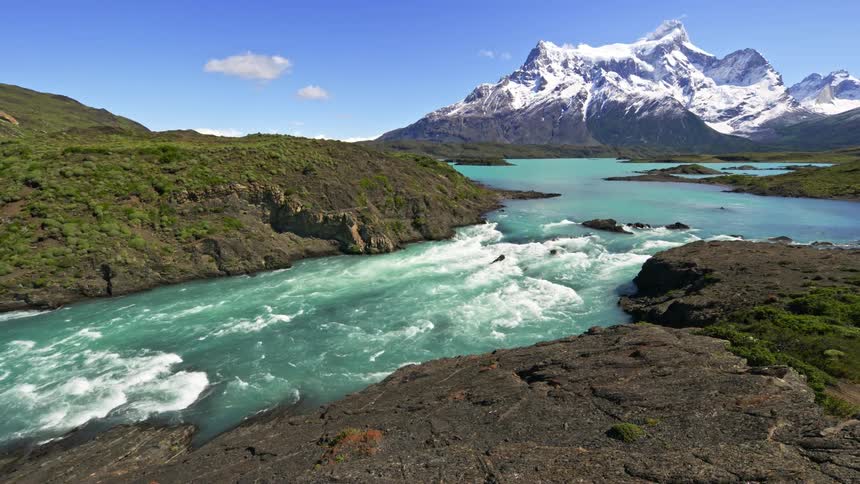 Blue river water flows past tall snow covered mountains and down rapids