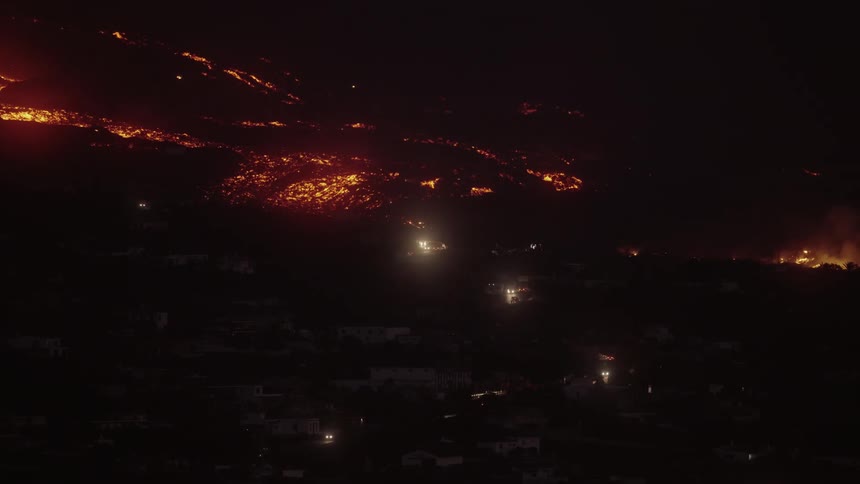 Lava from a volcano is coming close to a city that is dark and many cars are driving down the street