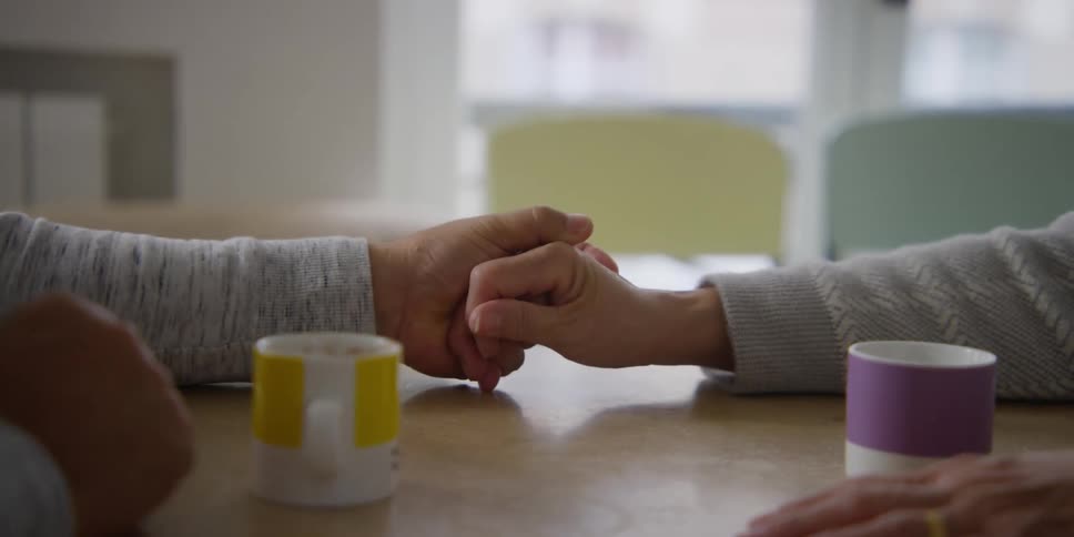Two people hold hands that are on a table and there are two colorful mugs