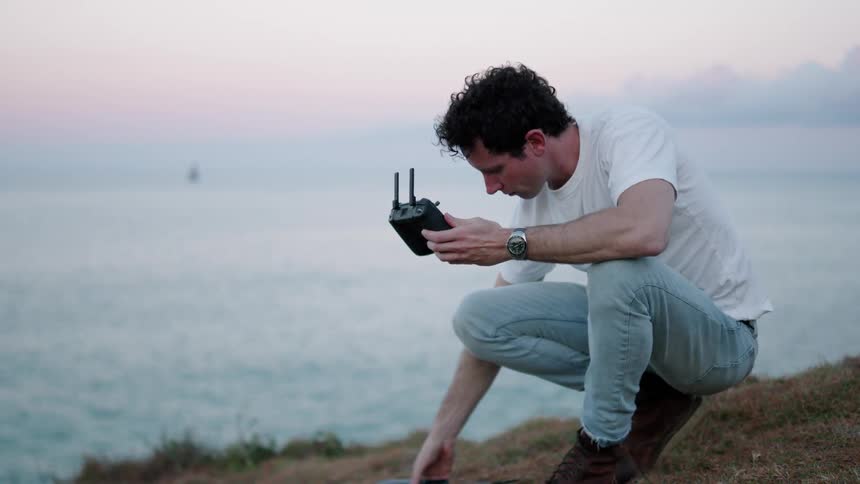 A man is crouched near the sea and looking at a controller in his hands