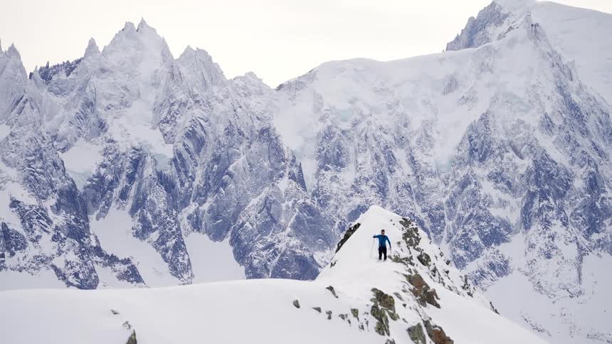 Male skier slowly walking in deep snow on mountain peak with ice covered mountains and bright white sky in the background