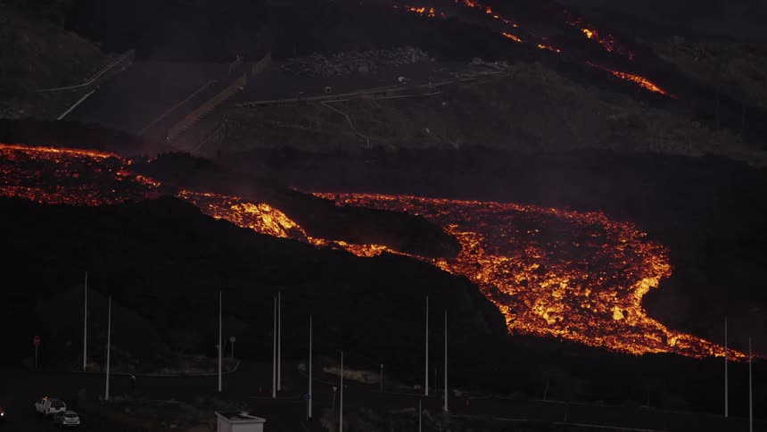 Large river of lava flows down a hillside destroying homes while an emergency responder surveys the road by a vehicle with flashing yellow lights nearby