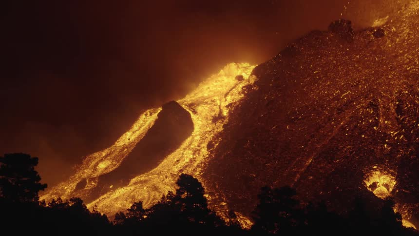 Volcano erupts with a fountain shooting up and a river of orange glowing lava flowing down the hillside showered in gold sparksat night in slow motion