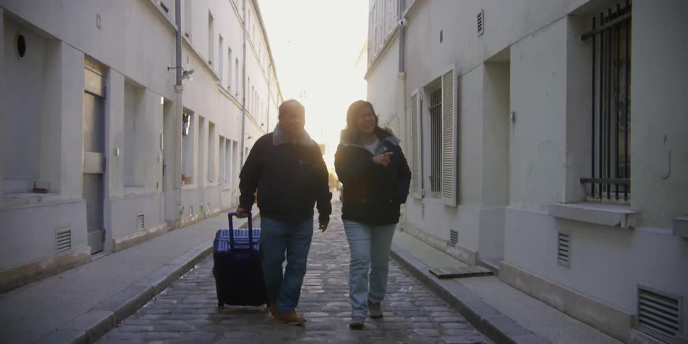An older couple walks with a suitcase through an alley