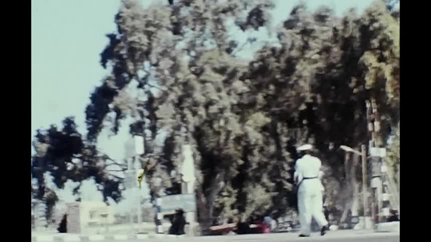 A person in white uniform standing on a sidewalk near a street cauhgt on vintage film