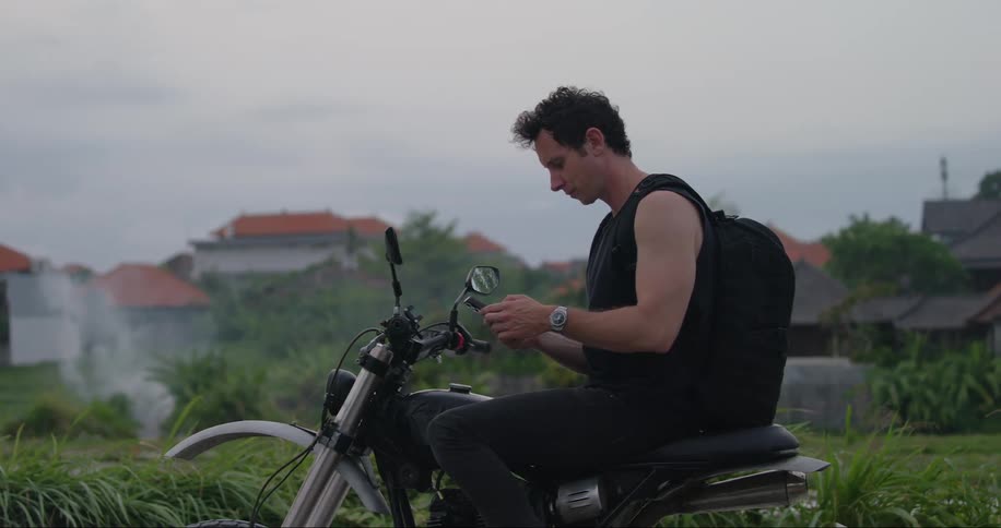 Man sitting on parked motorcycle in residential area looks around before accessing smartphone