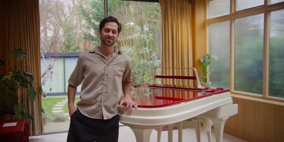 A smiling man stands next to a piano in a living room