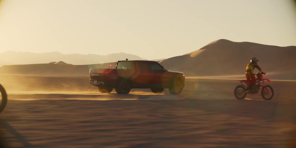 Two motorcycles and an suv car riding in a desert with several dunes and one of the motorcycles pushes its tire upwards
