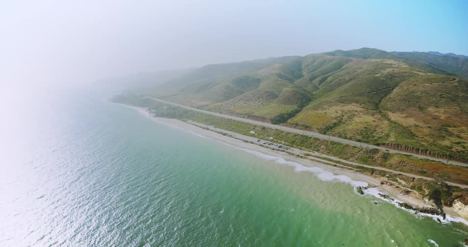 An aerial view of a beach with a large sea and then a highway with cars passing by