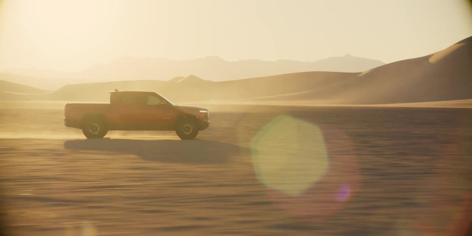 A truck driving in a desert and throwing a lot of sand behind it and there is strong sunlight in the background