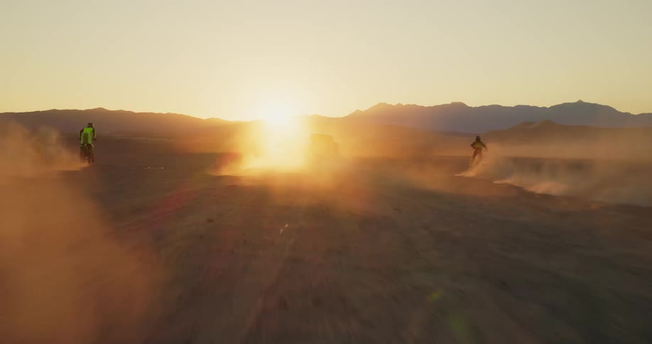 Pickup truck and two motorcycles speeding through the desert sands with the sun on the horizon