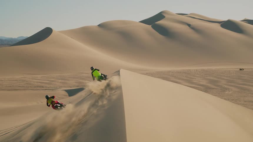 Two motorcyclists ride up and down side of large desert dune