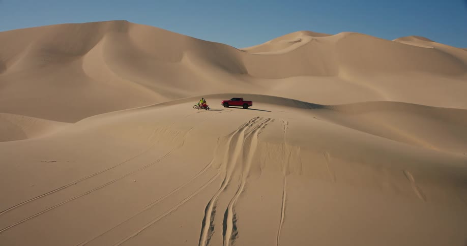 Motorcycles and a pickup truck park on top of a sand dune in the deserttion