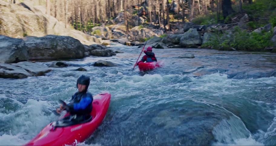 Two people are kayaking down a rapid river surrounded by rocks and trees