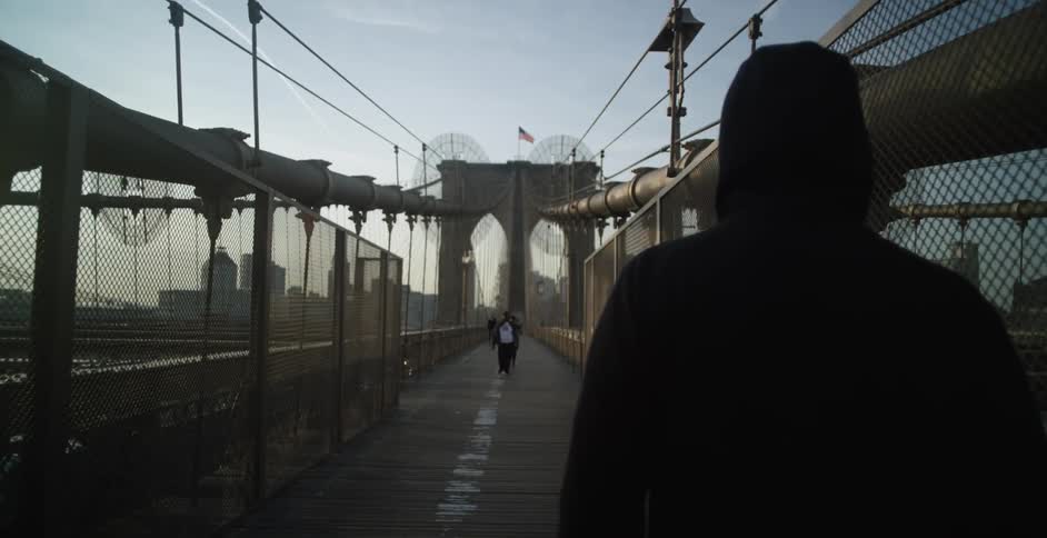 A man wearing a hoodie is walking over a pedestrian bridge in a city