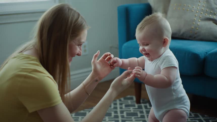 A mother is holding the hands of her baby who is standing on the floor and they are both smiling
