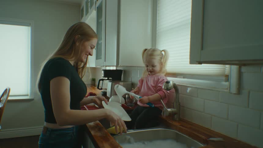 A little blonde girl inside the sink helping her mother wash dishes and they are both happy playing