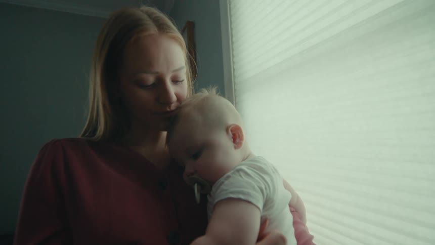 A young blonde mother is kissing her babys head which is on her lap