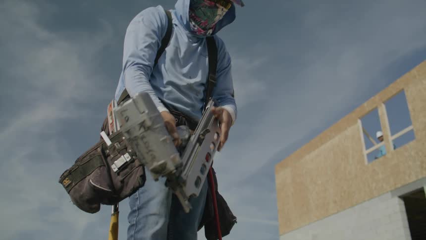 Construction worker nailing a wood frame laying on the ground with an air compressed nail gun and an unfinished building and construction materials in the background