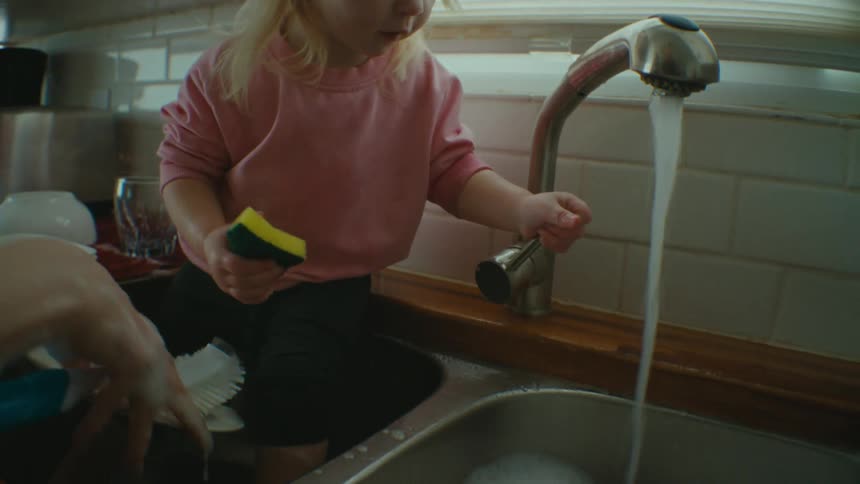 A little girl playing with the tap holding a dish sponge