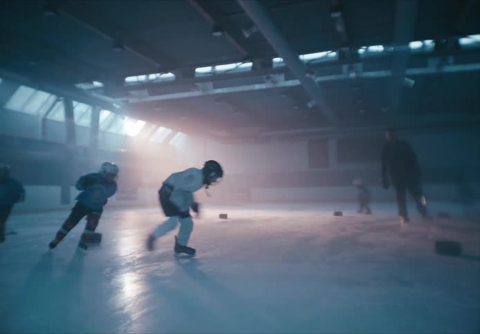 Youth hockey players practice ice skating in a circle around a coach on an indoor ice rink with the sunrise shining through in the background
